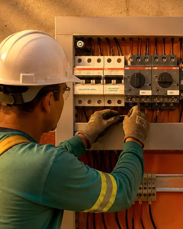 Técnico com capacete e luvas ajustando um painel elétrico de comando